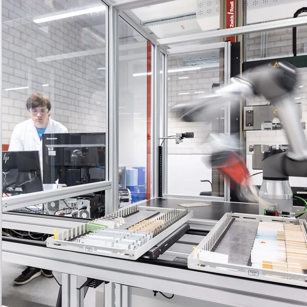 Lab technician Martin Altrock is working on a computer while sitting next to a lab robot.
