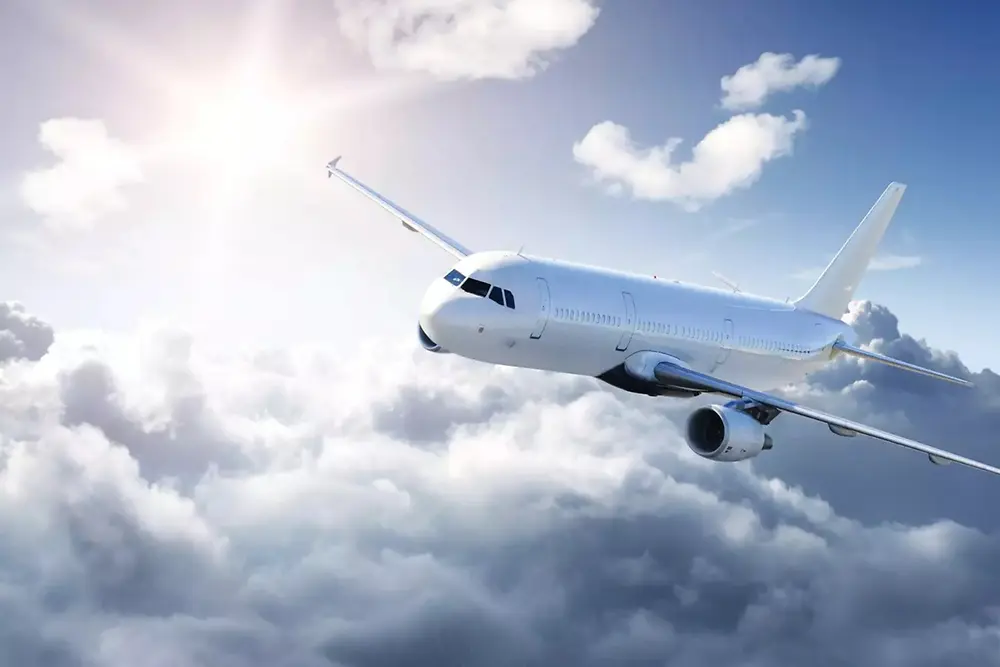 A white passenger airplane flying above soft clouds in a bright sky, with sunlight shining from the upper left.