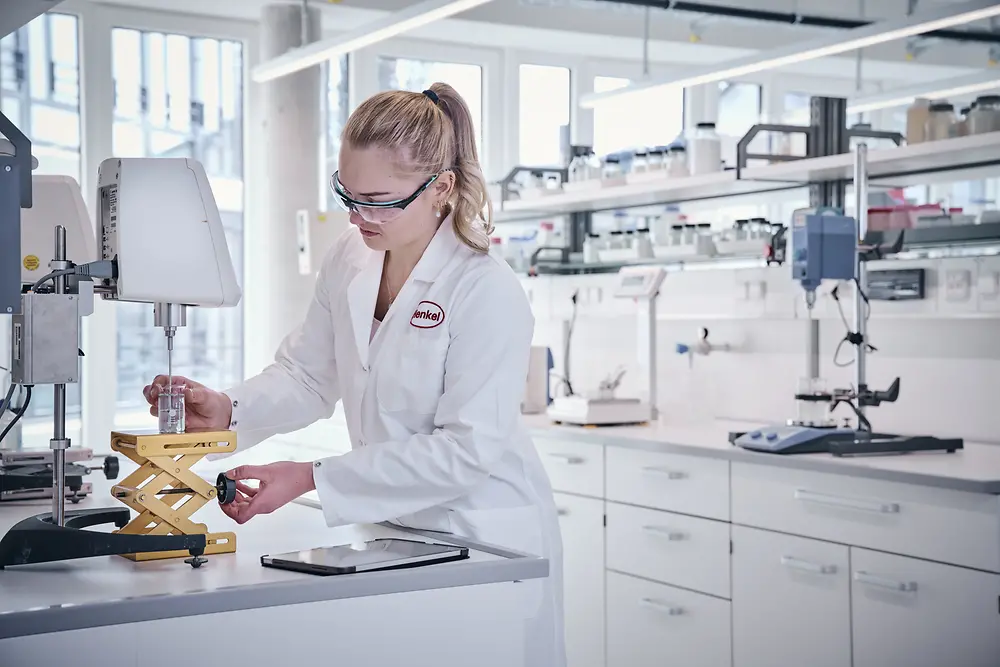 A Henkel researcher wearing a white lab coat and safety glasses is working in the lab. In the background, laboratory equipment, shelves, and large windows are visible.