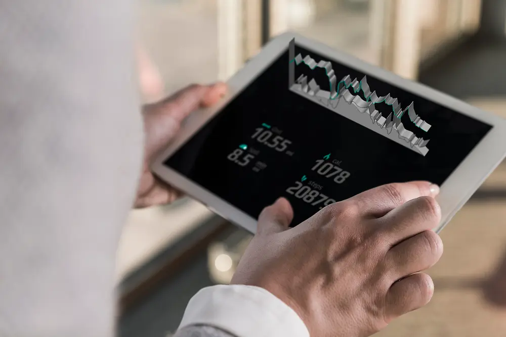 A person holding a tablet displaying digital charts and numeric data, viewed from over the shoulder in a bright indoor setting.